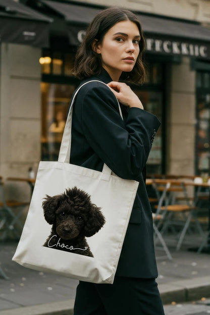 Woman carrying a natural cotton tote bag featuring a hyper-realistic portrait of a black toy poodle named Choco. Custom pet tote bag by JupiterStarCo, photographed outdoors in a stylish urban setting.