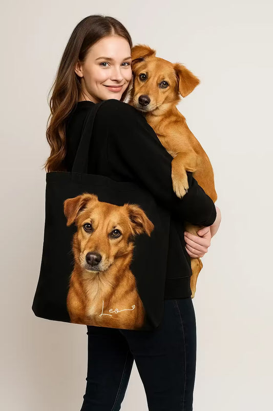 Woman holding a small brown dog while carrying a black tote bag featuring a hyper-realistic portrait of the same dog with the name "Leo" printed on it. Custom pet tote bag by JupiterStarCo.